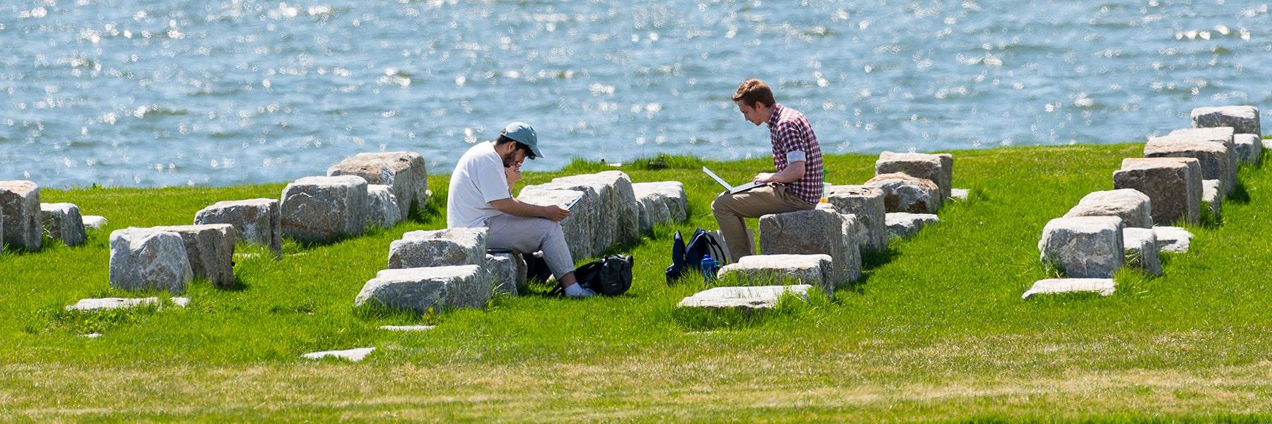 Students sit on stone benches by water on campus center lawn and work on laptops.